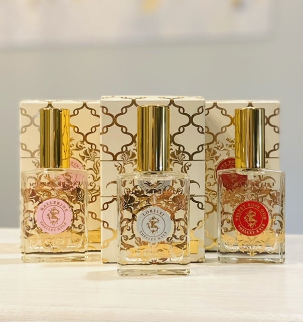 Three bottles of perfume lined up in a row with a box behind each clear glass rectangle bottle with gold lid
