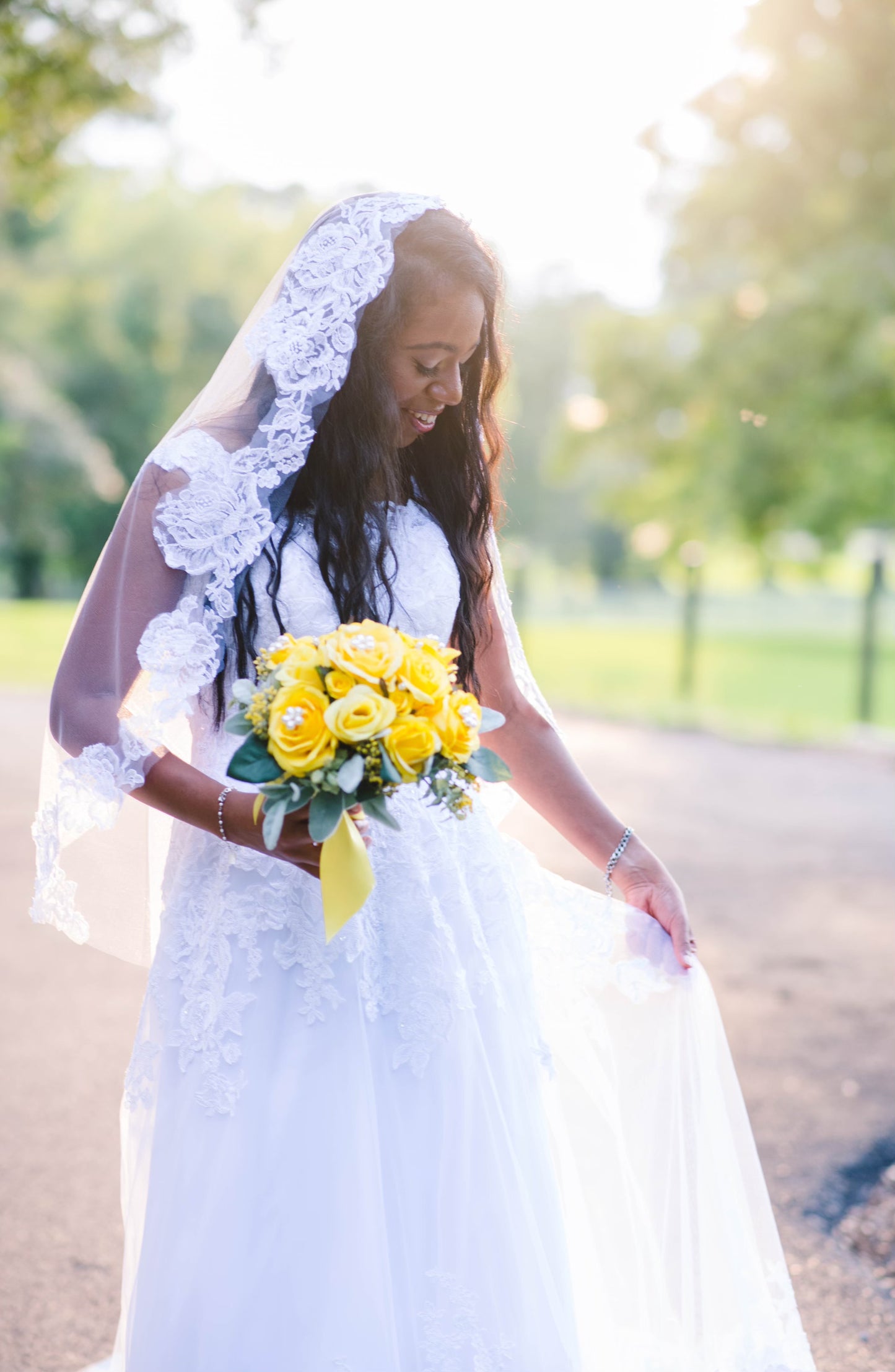A bride wearing a lace bridal veil with a metal comb, looking down at her dress with a bouquet of yellow roses.