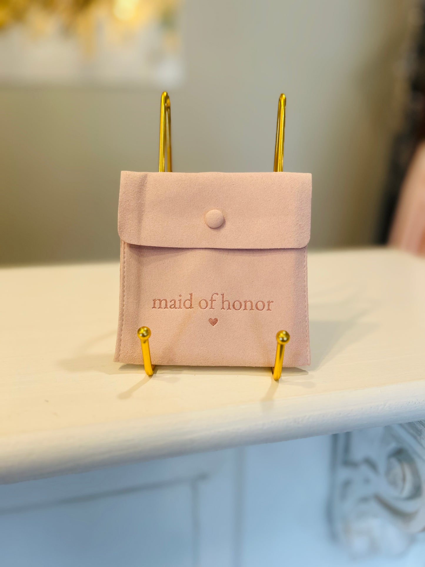 Pink pouch with 'maid of honor' text on a stand against a blurred background