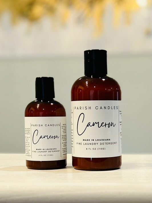 Two bottles of Parish Candles fine laundry detergent, one smaller and one larger, with dark caps and labels displaying the brand and scent 'Cameron'.