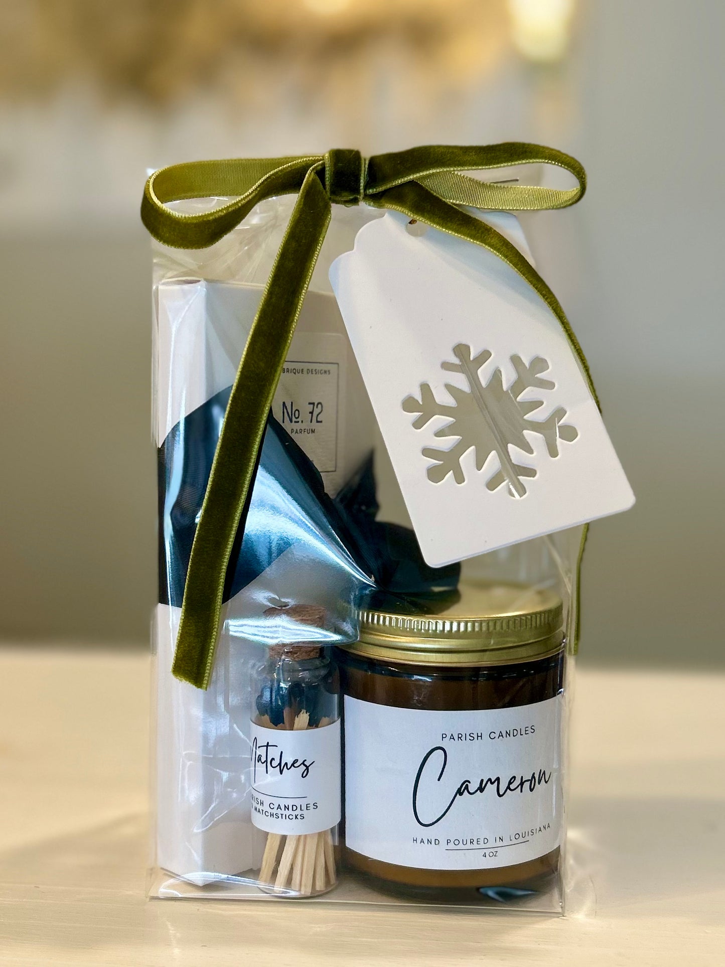 A gift set containing a clear bag with a green ribbon, a black and white spray bottle labeled 'No. 72 Perfume', a Cameron 4 ounce candle, and a set of matches, all tied together with a green bow.