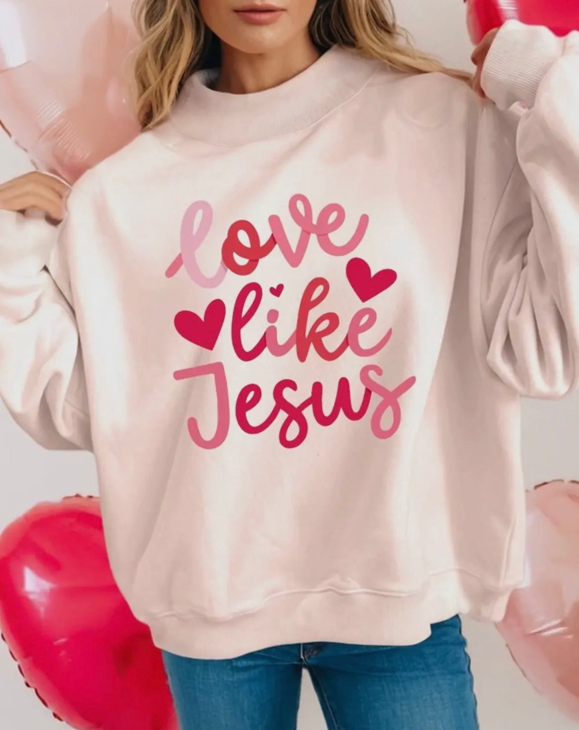 Person wearing a light pink sweatshirt with 'Love like Jesus' text and hearts, standing against a white background.