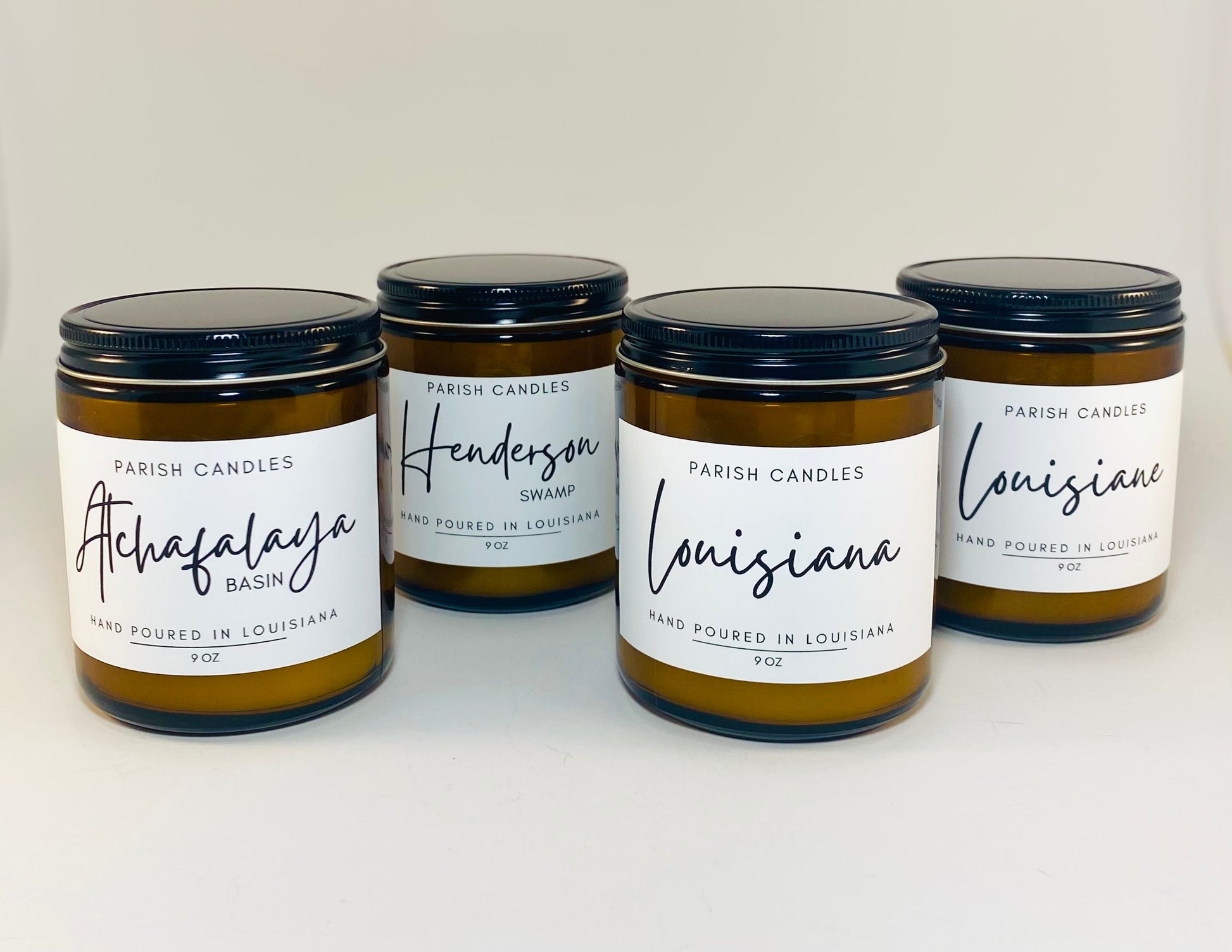 4 ounce candle in a horizontal row in brown jar and black lids with labels named, Atchafalaya Basin, Henderson Swam, Louisiana and Louisiane 