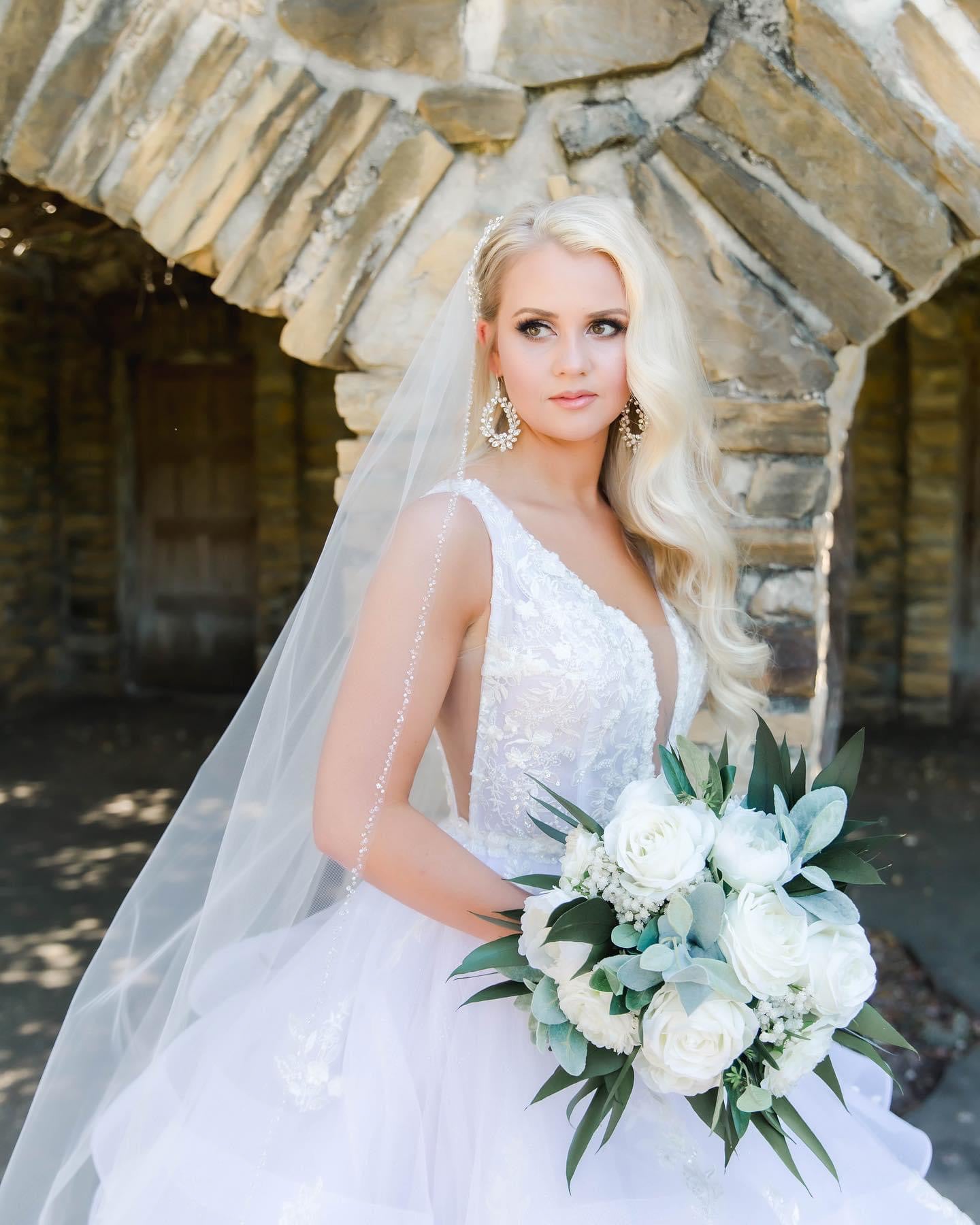 A woman wearing a white beaded bridal veil with a metal comb, standing in a stone-walled outdoor setting holding a bouquet of white flowers.