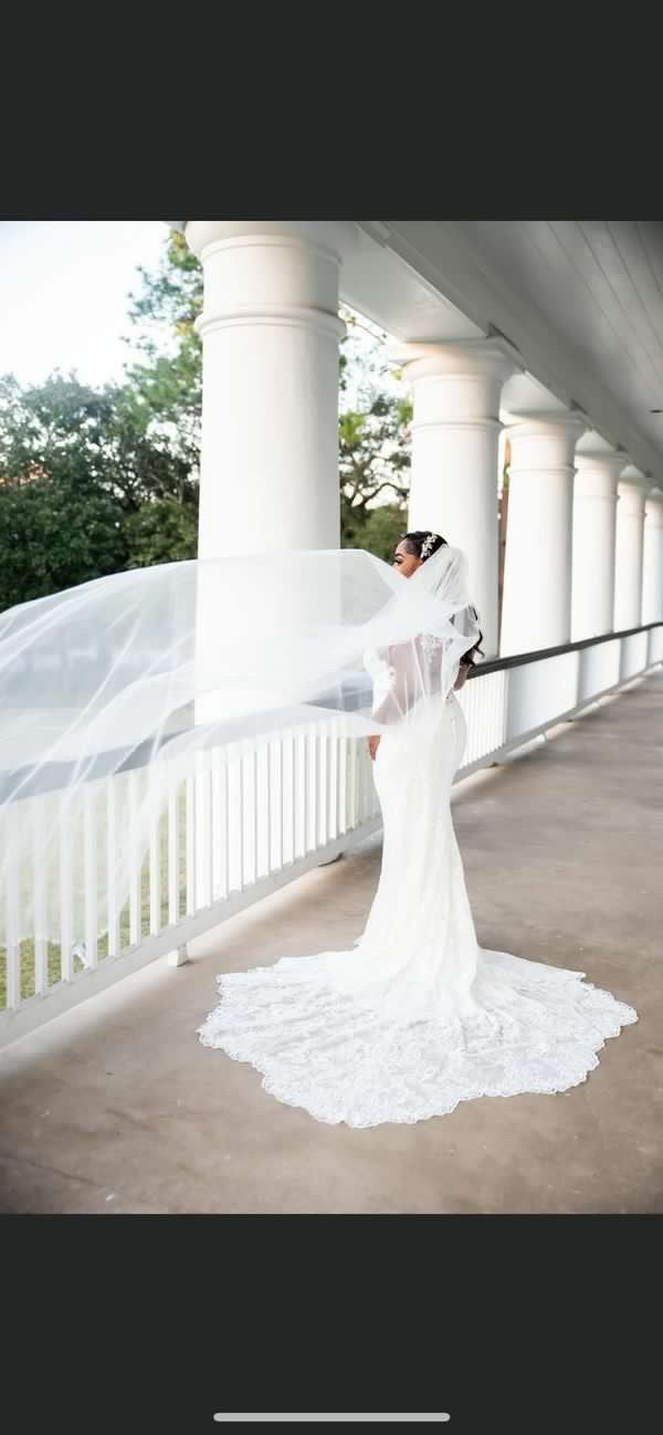 A bride wearing a custom beaded bridal veil with a metal comb, standing near a columned structure with a view of greenery in the background.