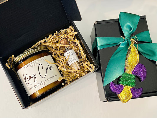 A Mardi Gras themed candle gift set with a 9 oz Parish Candles candle in a glass bottle with a cork stopper, a black gift box with green ribbon and a decorative purple and yellow ornament.