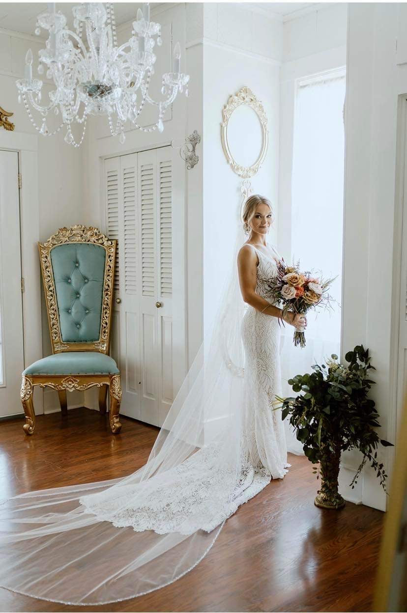 A bride wearing a custom beaded bridal veil with a metal comb, standing near a window and blue chair.